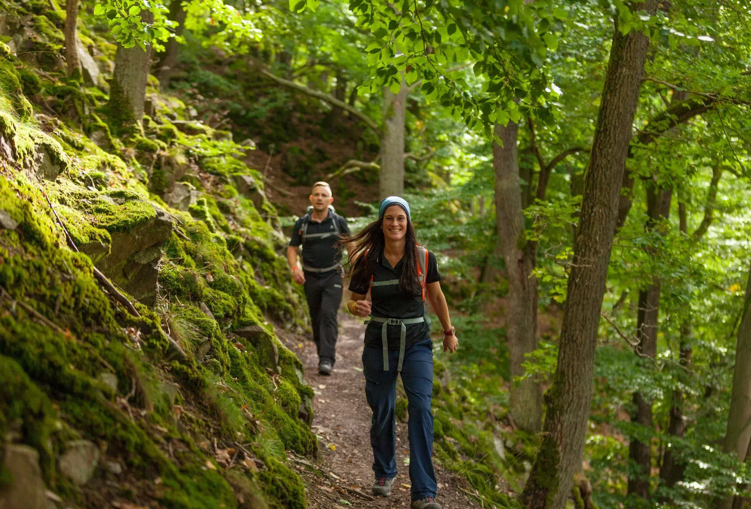 Ferienhaus Kellerwald Zwei Menschen wandern durch einen grünen, bewaldeten Pfad zwischen Bäumen und Moos Ferienhaus Edersee, Zertifizierung als „Qualitätsgastgeber – wanderbares deutschland“